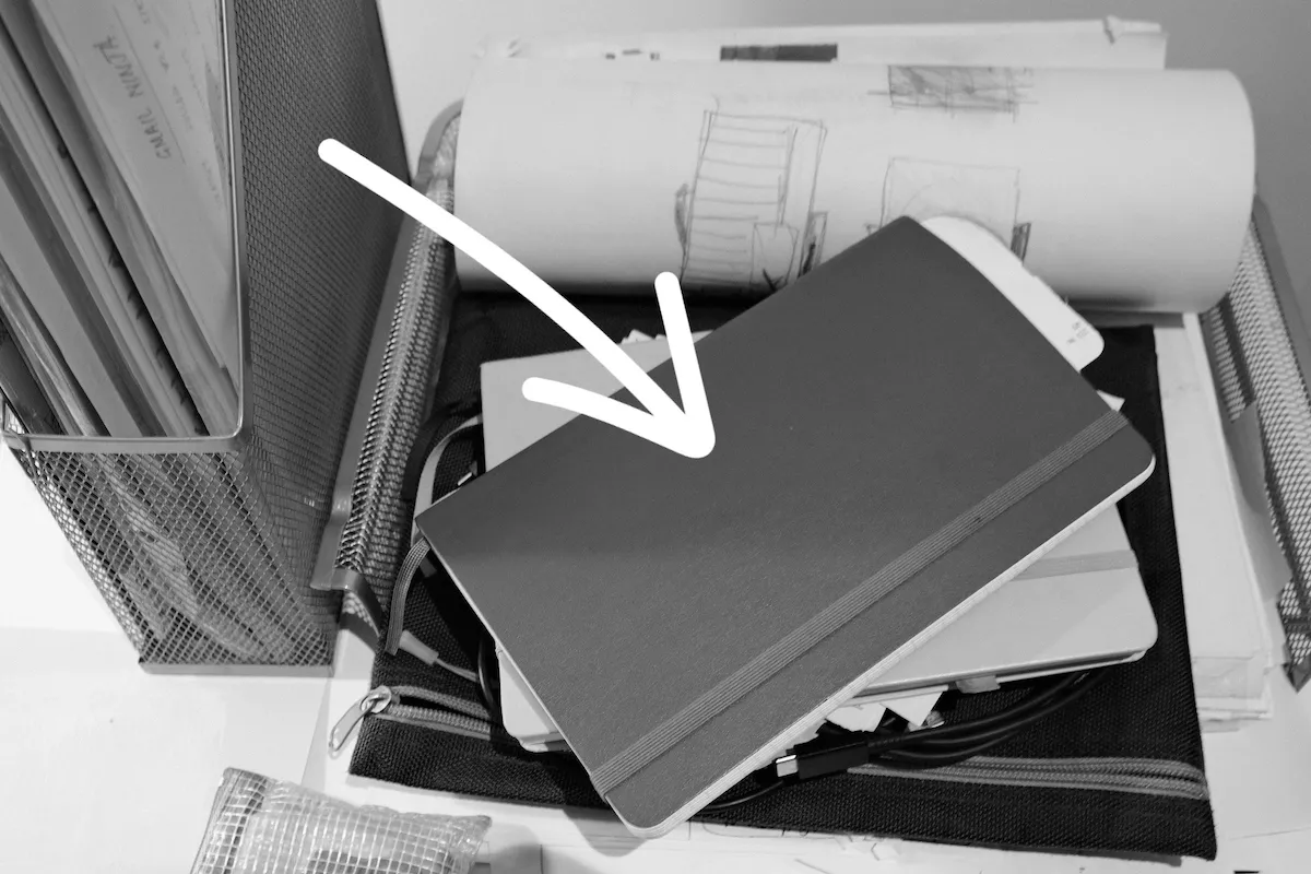 A black and white image of a notebook sitting in a full In-tray. It sits on top of aopther notebook, cables, a zipped foli, paper, and more.